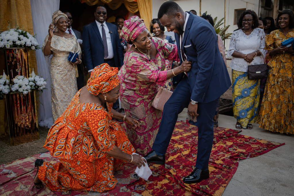 Noivo em fato azul é recebido de forma divertida por tias em trajes de panos africanos durante uma celebração tradicional angolana ao ar livre, enquanto familiares observam e riem.
